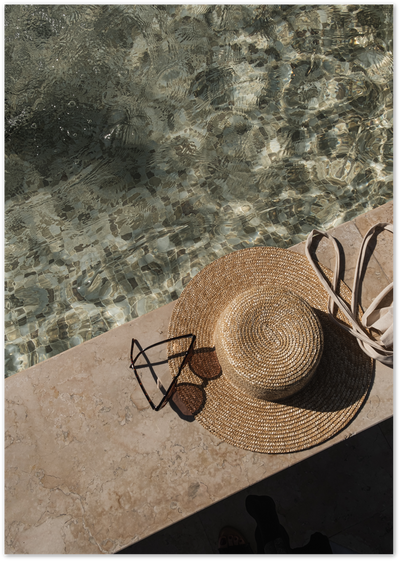 Straw hat, sunglasses and tote bag on marble poolside tiles beside rippling water